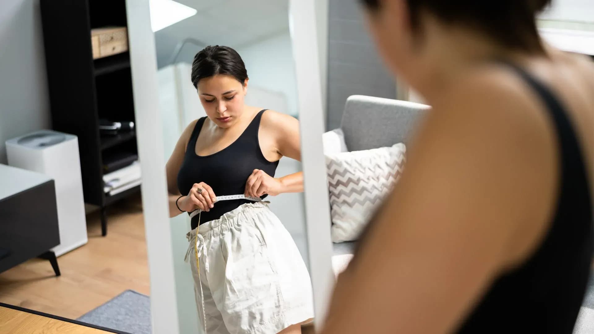 Woman measuring her waist with a tape measure while looking in a mirror, wearing a black tank top and light-colored shorts, in a modern home setting.