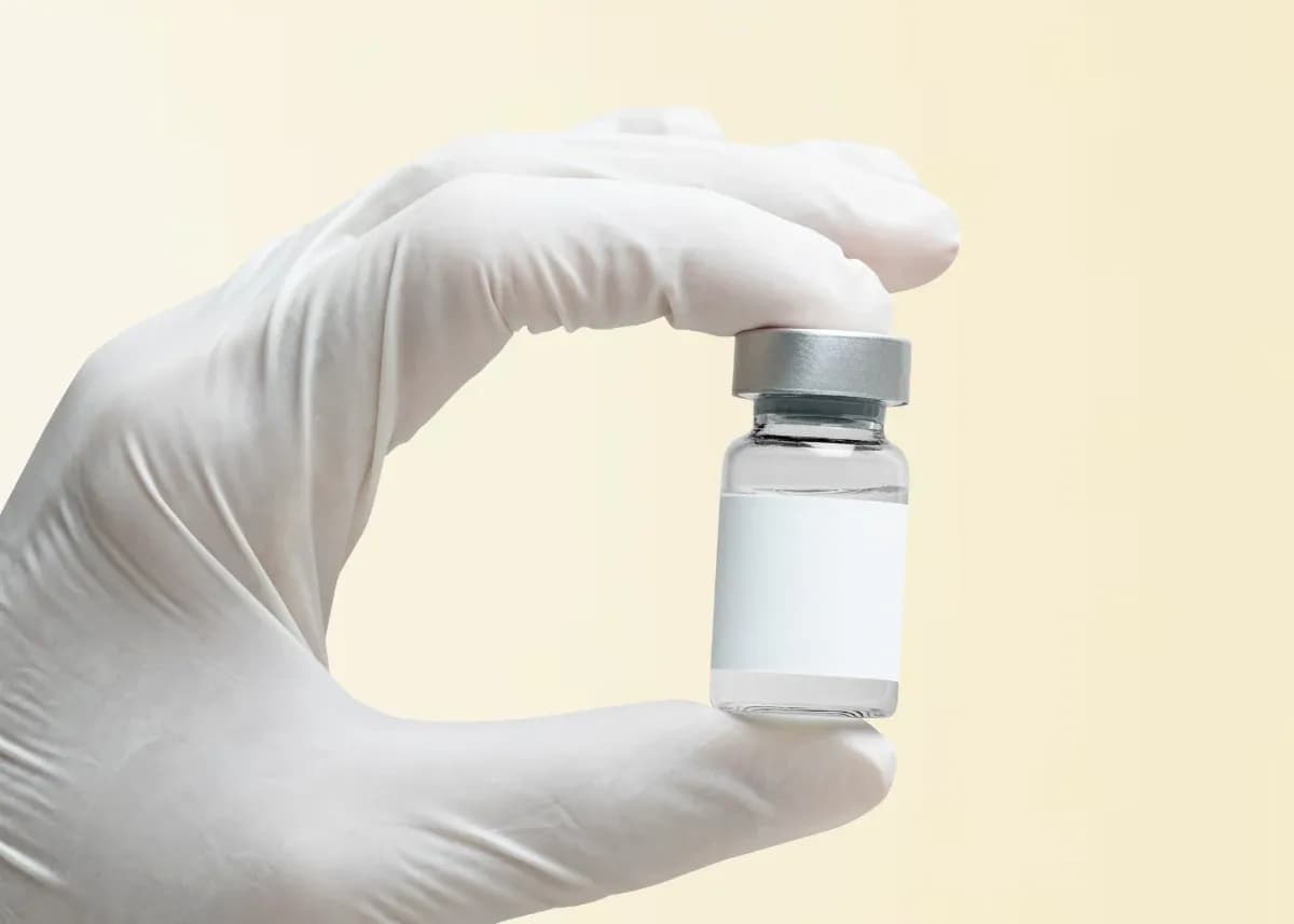 A gloved hand holding a small clear glass vial with a blank white label and a silver cap.