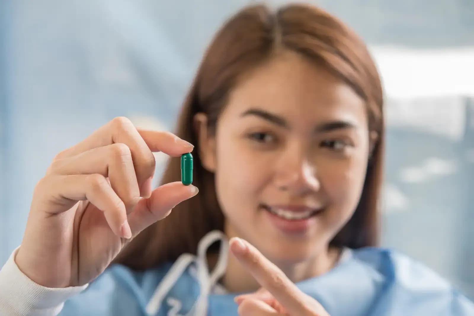 A smiling young woman in medical scrubs points at a capsule in her hand
